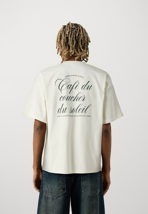 Person med blonde twists iført hvid T-shirt med fransk tekst "Café du coucher du soleil" og løse mørke denimjeans, stående mod en ensfarvet baggrund.