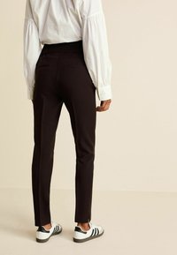 Zwarte op maat gemaakte broek met een slim fit en zijsplitten, gecombineerd met een witte blouse met lange mouwen en sportieve witte schoenen met groene accenten.