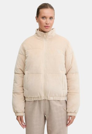 Donna che indossa una giacca imbottita beige con zip e collo alto, abbinata a pantaloni beige con motivo, in piedi davanti a uno sfondo semplice.