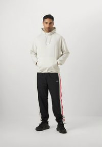 Light beige hoodie with front pocket and drawstrings, paired with black pants featuring red and white side stripes. Black sneakers complete the outfit.
