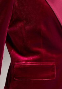 Dieprode fluwelen blazer met een slank ontwerp, een gladde textuur en een zak aan de linkerkant nabij de taille. Subtiele glans verbetert het uiterlijk.