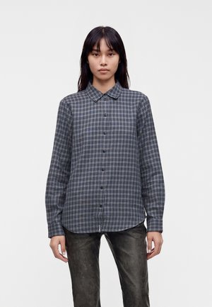 Jeune femme portant une chemise à carreaux gris foncé boutonnée et un pantalon noir texturé, debout devant un fond clair uni.