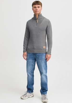 Grijze gebreide pullover met een hoge kraag en geribbelde accenten, voorzien van een halve rits en een leren logo-label aan de onderzijde. Draagt het met een blauwe jeans.