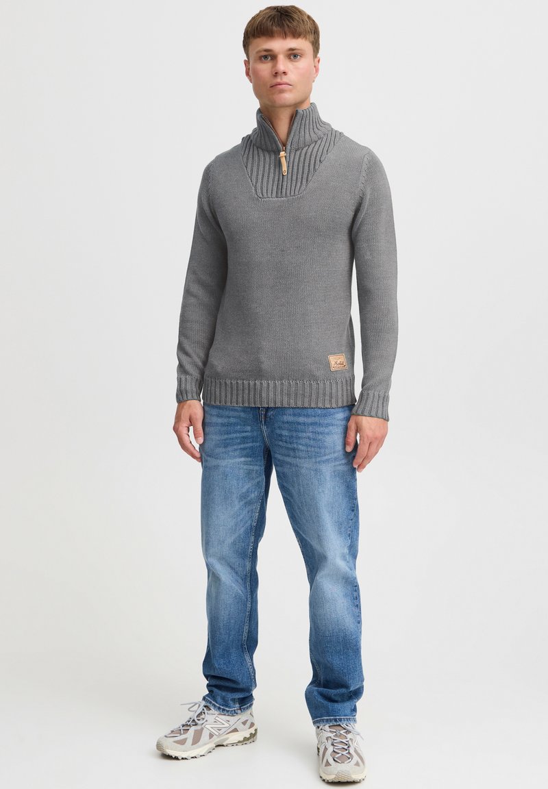 Pullover grigio a maglia con colletto alto e dettagli a coste, dotato di una cerniera a metà e un patch in pelle con logo sul lato inferiore. Indossato con jeans blu.