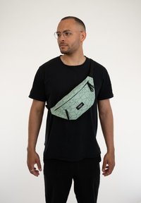 Marsupio verde chiaro con motivo geometrico, due tasche con zip, tracolla regolabile e dettaglio del logo. Indossato in stile crossbody.