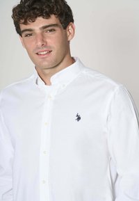 Camicia bianca button-up in cotone, con colletto a punta e piccolo logo di un cavallo blu navy sul lato sinistro del petto. Texture liscia, vestibilità su misura.