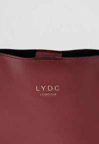 Primo piano di una borsa in pelle bordeaux con logo dorato "LYDC London" e bordo superiore visibile con fodera interna nera.