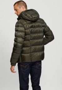 Puffer jacket in dark green with a quilted design, featuring a hood and ribbed cuffs. Fabric has a smooth, shiny texture and logo detail on sleeve.
