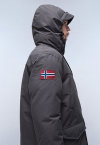 Grå isolerad jacka med huva, framfickor och en norsk flagga-patch på vänster ärm. Slät textur och praktisk design.