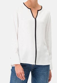 Blouse blanche à manches longues avec un bord noir, dotée d'une patte de boutonnage partielle et d'un col en V, fabriquée en tissu lisse.