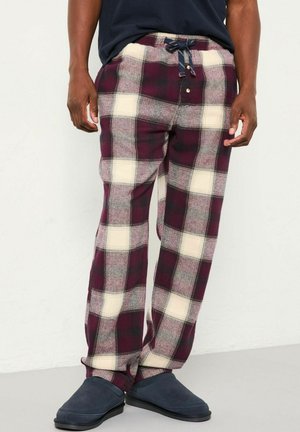 Man wearing burgundy, cream, and gray plaid pajama pants with navy slippers, standing against a plain white wall.