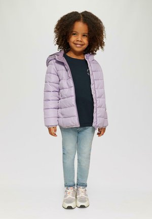 Lächelndes junges Mädchen mit lockigem Haar, das eine helllila Daunenjacke, ein schwarzes T-Shirt, hellblaue Jeans und pink-weiße Turnschuhe trägt.