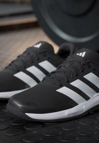 Chaussures de sport Adidas noires avec un logo blanc à trois bandes, placées sur un sol de gym en caoutchouc texturé avec une plaque de poids en arrière-plan.