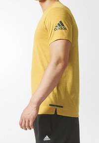 Camiseta atlética amarilla con tejido texturizado, mangas cortas y logo de Adidas en verde oscuro en el hombro izquierdo. Abertura lateral en el dobladillo.