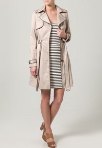 Beżowy trencz z dużym kołnierzem z wycięciem, dwurzędowymi guzikami i regulowanym paskiem. Nosi się go na dopasowaną sukienkę w czarno-białe paski.