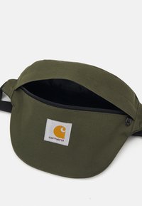 Marsupio di stoffa verde con cerniera nera, dotato di un'etichetta bianca con logo giallo. Texture liscia, design compatto, tracolla regolabile.