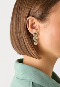 Orecchini a forma di cuore a tre livelli in oro e argento con accenti di cristallo, che presentano la parola "CUTE" sul cuore superiore, indossati da un modello.