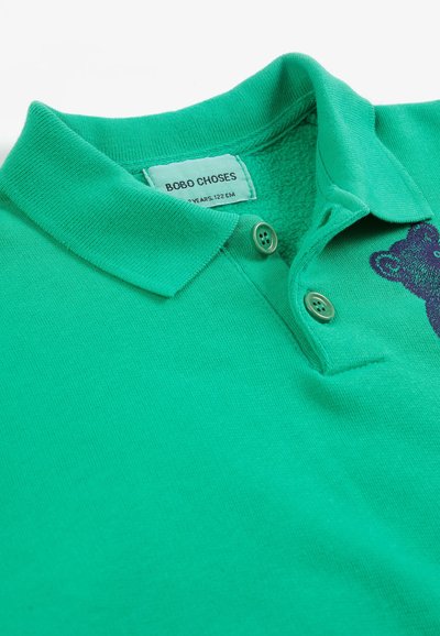 Polo vert pour enfant avec deux boutons, col et un petit motif d'ours près de l'encolure, étiquette de taille visible à l'intérieur du col.