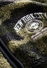 Zöld és fekete bolyhos kamuflázs fleecdzseki, hímzett "New York City" felirattal és kör alakú logóval, amelyen koponyadesign látható. Cipzáras elülső rész.