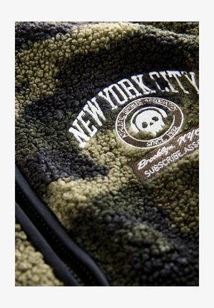 Vihreä ja musta kamuflaažile fleece-takki, jossa on brodeerattu "New York City" -teksti ja pyöreä logo, jossa on pääkallon kuvasto. Vetoketjukiinnitys edessä.
