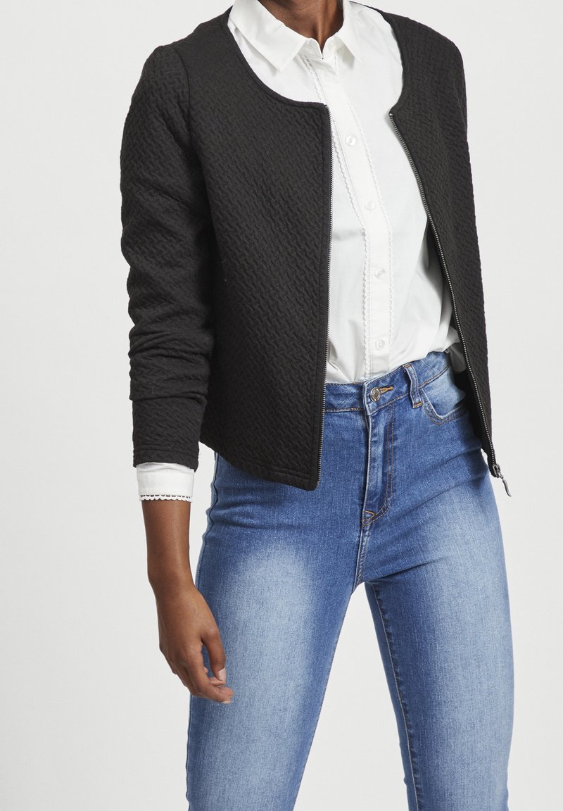 Personne portant une veste noire texturée zippée par-dessus une chemise blanche boutonnée, associée à un jean bleu clair ajusté, sur un fond uni.