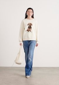Creme sweatshirt med et trykt brunt hundegraphic i midten, parret med blå denimbukser og en creme totebag. Struktureret stof, afslappet pasform.