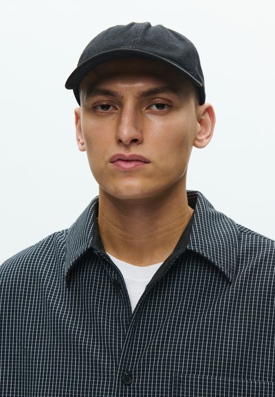 Μαύρο καπέλο του μπέιζμπολ, καρό ασπρόμαυρο πουκάμισο με γιακά που κουμπώνει. Λείο ύφασμα, ορατή ραφή, φορεμένο άνετα.