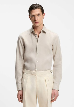 Mann trägt ein beige langärmeliges, geknöpftes Hemd, das in hoch taillierte, cremefarbene, plissierte Hose mit seitlichen Verstellriemen gesteckt ist.