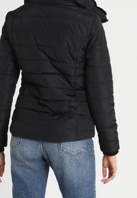 Svart puffjacka med horisontella sömmar, hög krage och figurnära passform. Matchad med ljusblå jeans.