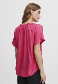 Top rosa a maniche corte con un motivo testurizzato, scollatura arricciata e orlo curvo, indossato con pantaloni neri.