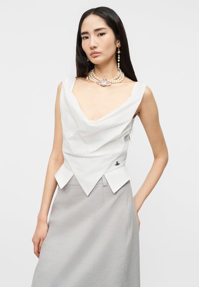 Γυναίκα με μακριά μαύρα μαλλιά φοράει άσπρο αμάνικο τοπ με πτυχώσεις, φούστα ανοιχτού γκρι και κολιέ από πέρλες με αντίστοιχα κρεμαστά σκουλαρίκια.