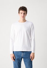 Hvit langermet t-skjorte laget av bomull med rund hals og et lite merke på brystet; kombinert med blå denim jeans.