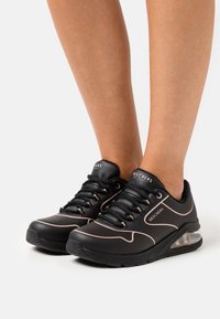 Black Skechers sneakers with a smooth texture, featuring light pink accents, a sturdy round toe, and a transparent air cushion sole.