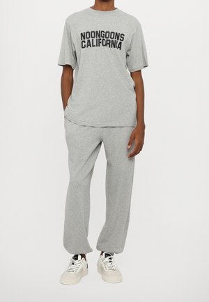 Persoon in een grijze oversized t-shirt met korte mouwen met de tekst "Noongoons California" en bijpassende grijze sweatpants met witte sneakers.