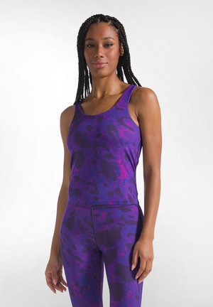 Mujer con el pelo trenzado que lleva una camiseta sin mangas morada y negra con estampado y leggings a juego, de pie contra un fondo claro.