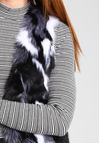Kamizelka z futra syntetycznego w czarno-białe łatki, noszona na pasiastym, czarno-białym, długim rękawie. Miękka tekstura, warstwowy fason, brak widocznych metalowych elementów.