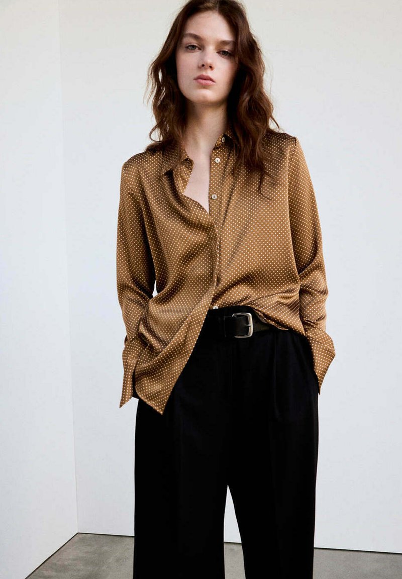 Jeune femme aux cheveux ondulés porte un chemisier marron à pois, rentré dans un pantalon noir taille haute avec une ceinture noire, debout contre un mur uni.