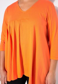 Oranges V-Ausschnitt-Oberteil mit Dreiviertelärmel, ausgestattet mit plissiertem Design, weicher Textur und lockerer Passform, die sanft über den Körper fällt.