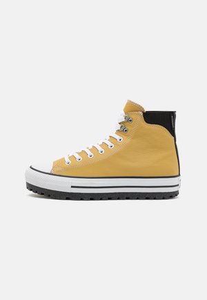 Żółty sneaker typu high-top z białymi sznurowadłami, czarną pętelką na pięcie, grubą białą podeszwą z czarnym paskiem oraz fakturowaną czarną podeszwą zewnętrzną.