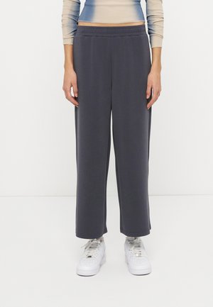 Persoană îmbrăcată cu pantaloni largi gri închis, pantofi sport albi și un top bej cu mâneci lungi, stând în fața unui fundal alb simplu.