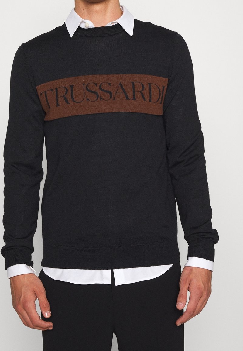 Homme portant un pull noir avec une bande horizontale marron et le mot « TRUSSARDI » sur la poitrine, par-dessus une chemise blanche boutonnée.