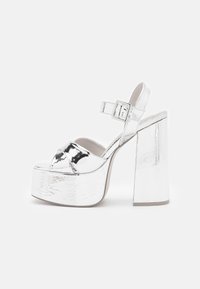 Stříbrné metalické platformové sandály s širokými křížovými pásky, páskem kolem kotníku se sponou a vysokým masivním podpatkem na bílém pozadí.