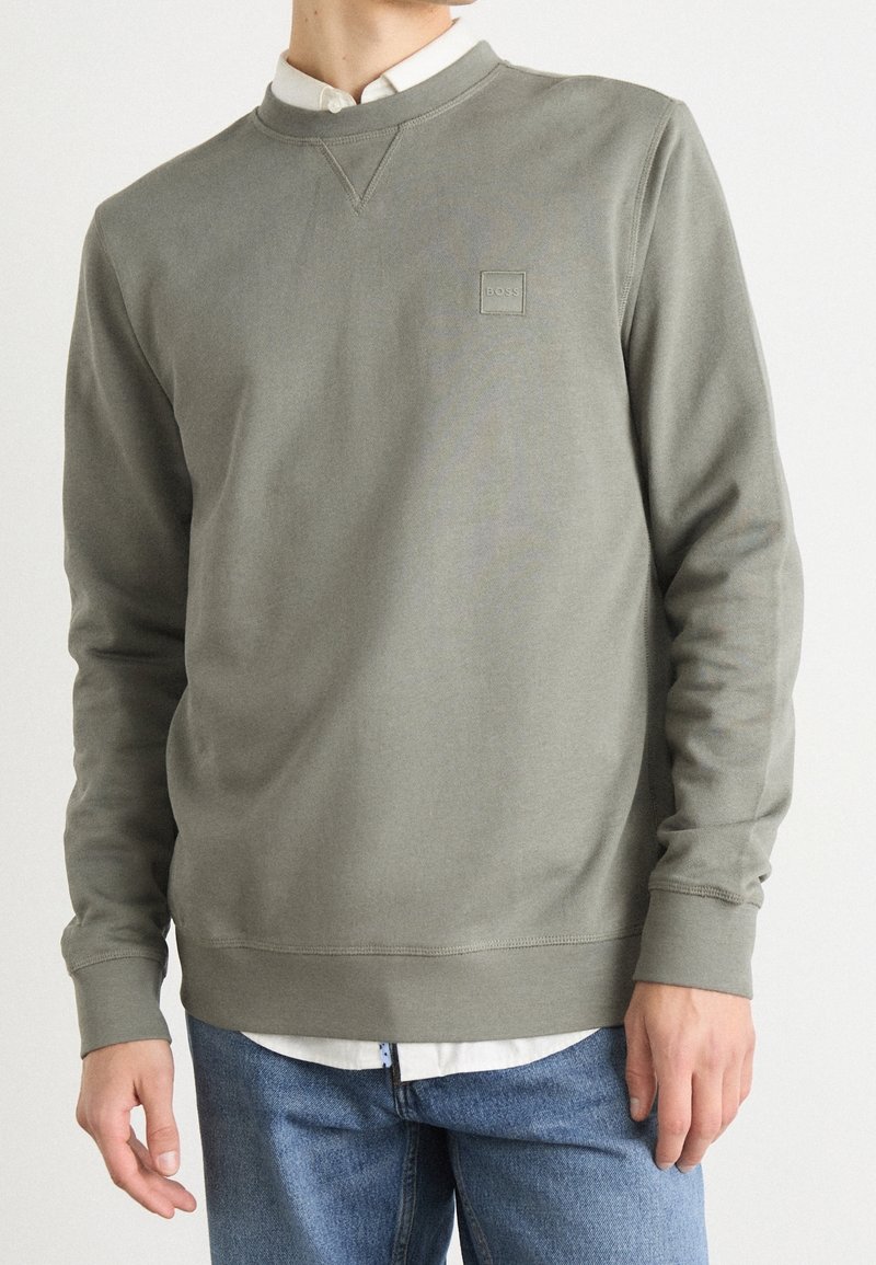 Personne portant un sweat-shirt vert olive clair avec le logo BOSS sur la poitrine, par-dessus une chemise blanche à col, associé à un jean bleu, coupé au niveau du cou et de la taille.