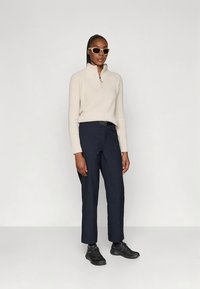 Haut zippé en polaire beige clair avec un col montant, associé à un pantalon droit bleu marine foncé et des chaussures de sport noires. Lunettes de soleil blanches épaisses.