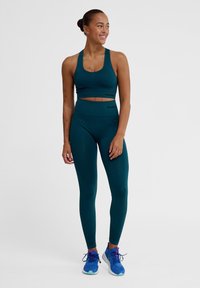 Zestaw odzieży sportowej w kolorze teal, składający się z krótkiego topu i legginsów z wysokim stanem, wykonany z gładkiego, elastycznego materiału. Wykończony detalami z logo.