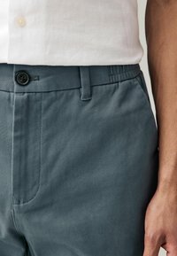 I pantaloni tessuti grigio-blu presentano una chiusura a bottone, tasche laterali e una cintura elastica. Il tessuto ha una texture liscia e una leggera lucentezza.