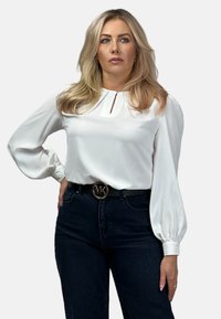 Biała satynowa bluzka z długimi, bufiastymi rękawami i detalem dekoltu w kształcie łezki. Zestawiona z ciemnoniebieskimi dżinsami i czarnym paskiem z złotym logo.