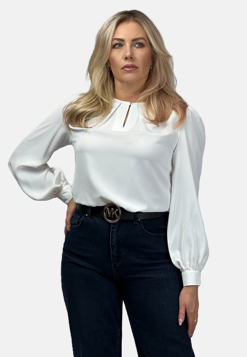 Biała satynowa bluzka z długimi, bufiastymi rękawami i detalem dekoltu w kształcie łezki. Zestawiona z ciemnoniebieskimi dżinsami i czarnym paskiem z złotym logo.