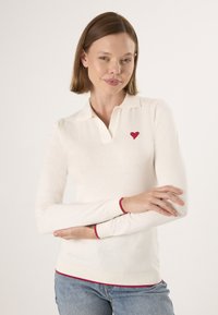 Witte lange mouwen polo met een klein rood hartembleem op de linkerborst en rode accenten bij de manchetten en de zoom. Zacht breimateriaal.