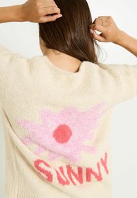Pull en beige avec une texture douce et duveteuse. Présente un grand motif floral rose et rouge avec le mot "SUNNY" en lettres majuscules au dos.